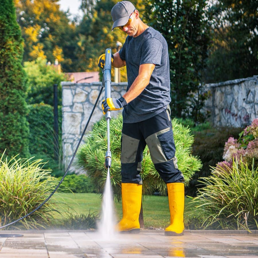 A technician power washing a paved patio, spray lifting dirt off the slabs